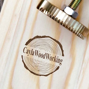 Puede incluir: Una tabla de madera con un sello de madera quemada que dice "Cayla WoodWorking" en un círculo alrededor de un diseño de anillo de árbol. También hay un tornillo de metal con cabeza de latón en la imagen.