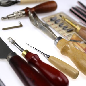 May include: A collection of leatherworking tools, including various knives, awls, and screwdrivers. The tools have wooden and metal handles, with some featuring red and natural wood tones. A variety of blades and cutting edges are visible, suggesting a focus on precision and detail.