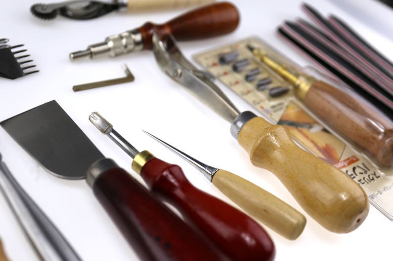 May include: A collection of leatherworking tools, including various knives, awls, and screwdrivers. The tools have wooden and metal handles, with some featuring red and natural wood tones. A variety of blades and cutting edges are visible, suggesting a focus on precision and detail.