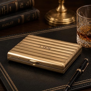May include: A gold-colored, rectangular case with horizontal ridges and the engraved initials "J. DOE." The case is on a black leather surface, next to a pen, a glass of amber liquid, and books. The case has a small clasp.