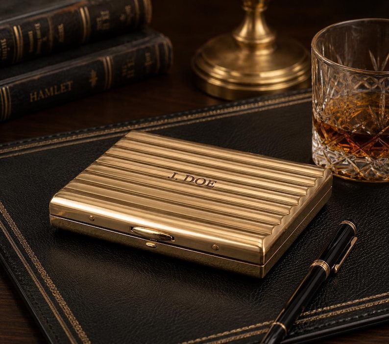 May include: A gold-colored, rectangular case with horizontal ridges and the engraved initials "J. DOE." The case is on a black leather surface, next to a pen, a glass of amber liquid, and books. The case has a small clasp.