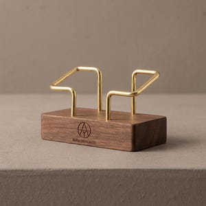 May include: A rectangular dark brown wooden base with two gold-coloured metal wire supports extending upwards. The front of the wooden base features a circular logo and the text AURA DESIGN CO. printed in black ink on the smooth wood surface.