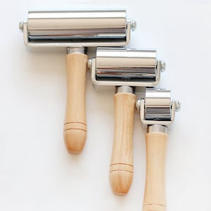 May include: Three metal hand rollers with wooden handles of varying sizes. The rollers have a shiny, silver finish and are attached to the wooden handles with a metal frame. The handles are light-colored wood. The tools are arranged diagonally against a white background.
