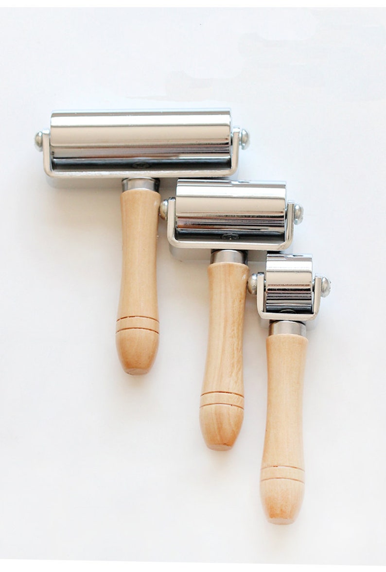 May include: Three metal hand rollers with wooden handles of varying sizes. The rollers have a shiny, silver finish and are attached to the wooden handles with a metal frame. The handles are light-colored wood. The tools are arranged diagonally against a white background.