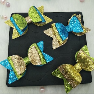 Peut inclure: Quatre nœuds en paillettes dans différentes combinaisons de couleurs. Les nœuds sont vert et bleu, or et bleu, or et vert, et bleu et or. Les nœuds sont sur une feuille de mousse noire avec des découpes.