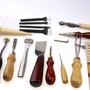 May include: A collection of leatherworking tools, including various knives, punches, and stitching tools. The tools have wooden and metal handles, and some are packaged in a clear plastic container. The tools are arranged on a white surface.