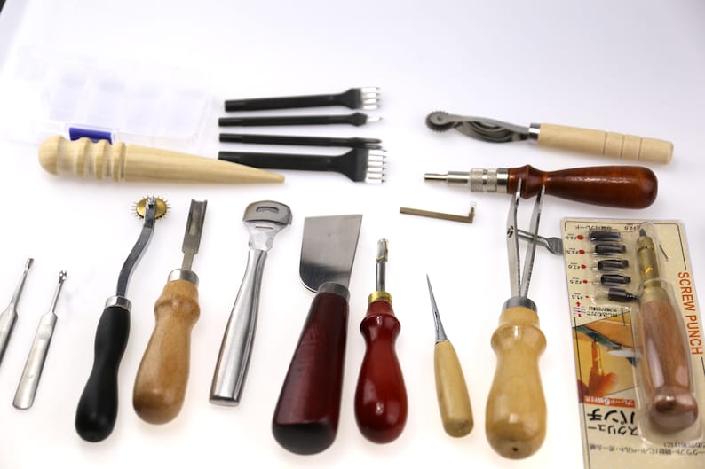 May include: A collection of leatherworking tools, including various knives, punches, and stitching tools. The tools have wooden and metal handles, and some are packaged in a clear plastic container. The tools are arranged on a white surface.