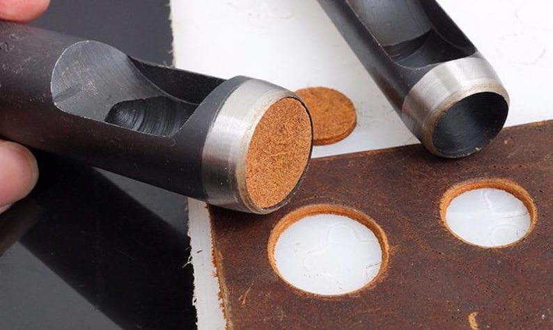May include: Two metal leather hole punch tools with a brown leather surface with three holes punched out. The tools have a silver metal rim and a brown leather pad on the end.