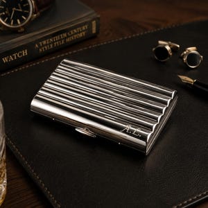 May include: A silver business card holder with a ribbed design and the initials "A.L." engraved on the front. The holder is on a black leather surface, with a watch, a book, cufflinks, a pen, and a glass of amber liquid in the background.