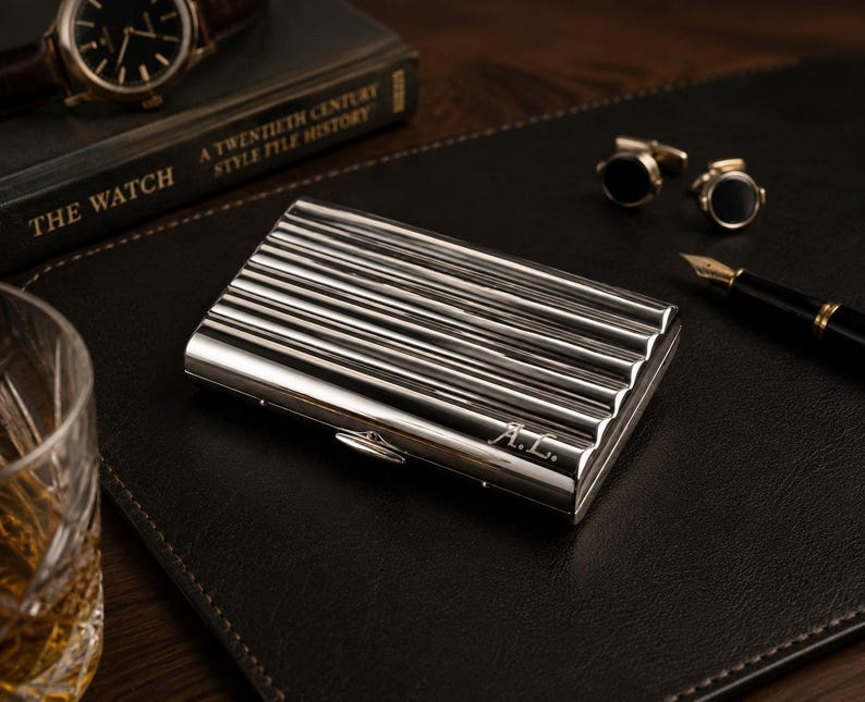 May include: A silver business card holder with a ribbed design and the initials "A.L." engraved on the front. The holder is on a black leather surface, with a watch, a book, cufflinks, a pen, and a glass of amber liquid in the background.