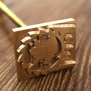 May include: A gold-colored metal branding iron with a square head and a long handle. The head has a detailed design of a gear with a circle in the center.