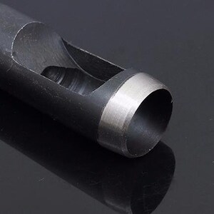May include: A close-up of a black metal hole punch with a silver rim around the cutting edge. The punch is designed to create round holes in materials like leather or paper.