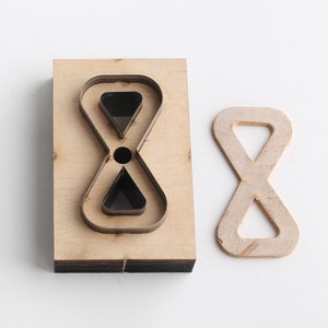 Pu&ograve; includere: Un punzone artigianale in legno con un ritaglio a forma di simbolo di infinito. Il punzone &egrave; su una superficie bianca, e un campione del ritaglio &egrave; accanto.