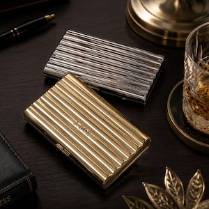 May include: Two metal business card holders, one silver and one gold, with a ribbed design. The gold holder is engraved with the initials "J. DOE". The holders are on a dark wooden surface, with a pen, a glass of whiskey, and a notepad in the background.