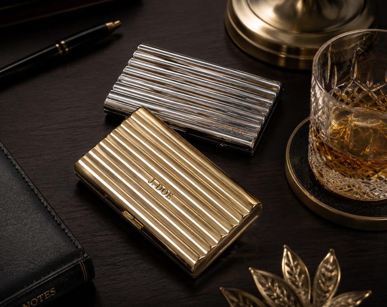 May include: Two metal business card holders, one silver and one gold, with a ribbed design. The gold holder is engraved with the initials "J. DOE". The holders are on a dark wooden surface, with a pen, a glass of whiskey, and a notepad in the background.