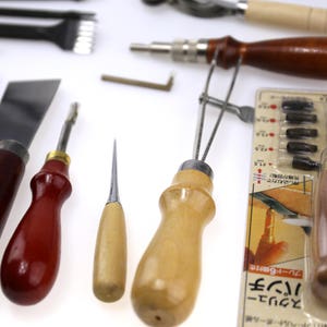 May include: Assortment of leatherworking tools, including a screw punch, awls, knives, and edge creasers. The tools have wooden and metal handles, with a screw punch package visible. The tools are arranged on a white surface.