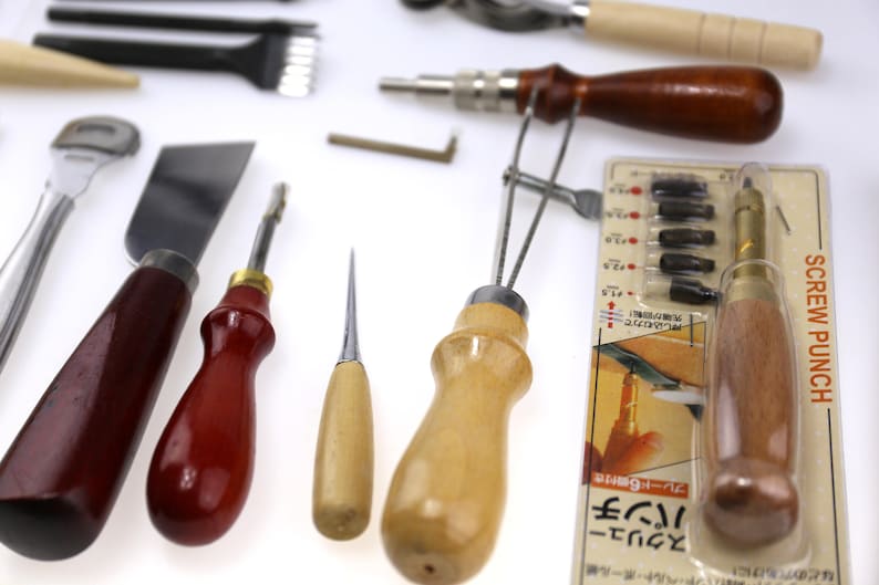 May include: Assortment of leatherworking tools, including a screw punch, awls, knives, and edge creasers. The tools have wooden and metal handles, with a screw punch package visible. The tools are arranged on a white surface.