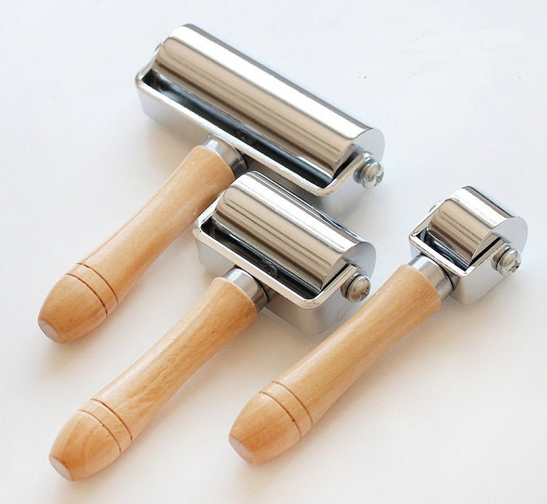 May include: Three metal and wood hand rollers of varying sizes are arranged on a white surface. Each roller has a silver-colored cylindrical roller attached to a rectangular metal frame. The handles are made of light-colored wood.