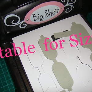 Puede incluir: Una hoja de papel blanca con varias formas recortadas. El papel est&aacute; colocado en una m&aacute;quina de troquelado negra con el texto "Big Shot" en ella. El texto "Suitable for Sizzix!" est&aacute; escrito en letras rosas en el papel.
