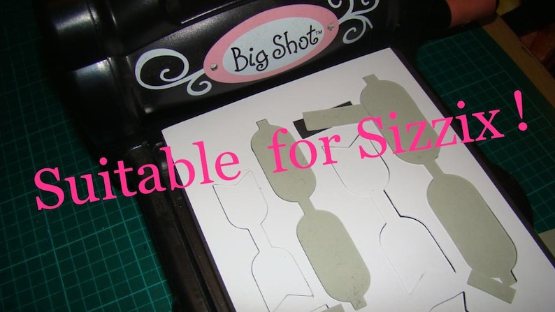 Puede incluir: Una hoja de papel blanca con varias formas recortadas. El papel est&aacute; colocado en una m&aacute;quina de troquelado negra con el texto "Big Shot" en ella. El texto "Suitable for Sizzix!" est&aacute; escrito en letras rosas en el papel.