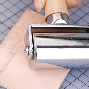 May include: A metal roller with a wooden handle is pressed onto two pieces of light brown leather. The roller is silver and shiny. The leather pieces are on a gray cutting mat with a grid pattern. The handle is light brown.