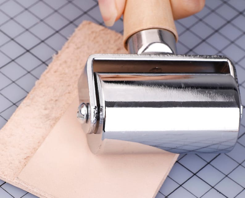 May include: A metal roller with a wooden handle is pressed onto two pieces of light brown leather. The roller is silver and shiny. The leather pieces are on a gray cutting mat with a grid pattern. The handle is light brown.