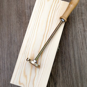 May include: A wooden handle attached to a metal rod with a toothed wheel at the end. The tool is lying on a light brown wooden surface.