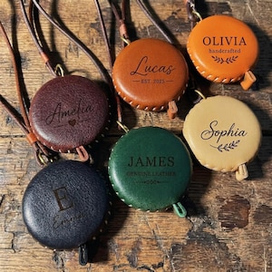 Op de afbeelding: Een verzameling gepersonaliseerde leren meetlinten in verschillende kleuren, waaronder bruin, oranje, groen en beige. Elk meetlint heeft een naam of initiaal, sommige met de tekst "handcrafted" of "genuine leather".