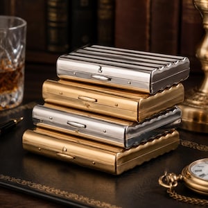 May include: A stack of four metal cigarette cases. Two are silver with a ribbed design, and two are gold-toned with a wavy edge. The cases are stacked on a dark leather surface, with a glass of amber liquid and a pocket watch in the background.