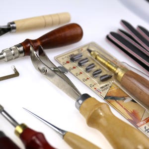 May include: Assortment of leatherworking tools, including a stitching groover, edge beveler, and various punches. The tools have wooden and metal handles, with a package of replacement blades and several black and red sanding sticks.