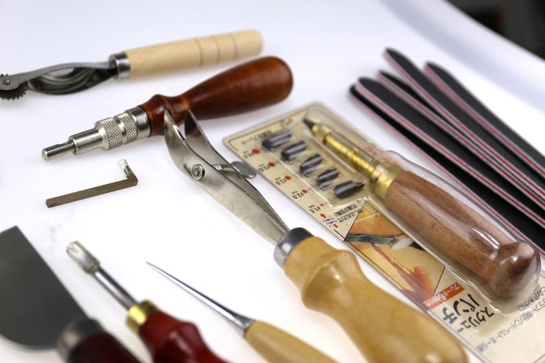May include: Assortment of leatherworking tools, including a stitching groover, edge beveler, and various punches. The tools have wooden and metal handles, with a package of replacement blades and several black and red sanding sticks.