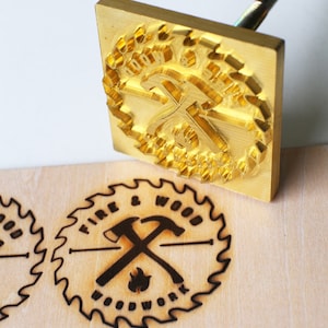 May include: A brass wood burning branding iron with a circular design featuring crossed hammers, a flame, and the text "Fire & Wood Woodwork".