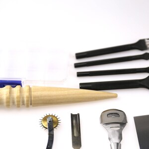 May include: A collection of leatherworking tools, including black punches with various prong configurations, a wooden handle awl, and a rotary stitch marker. The tools are arranged on a white surface, suggesting a craft or hobby setting.