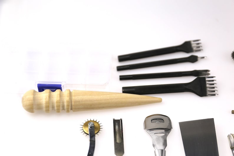 May include: A collection of leatherworking tools, including black punches with various prong configurations, a wooden handle awl, and a rotary stitch marker. The tools are arranged on a white surface, suggesting a craft or hobby setting.