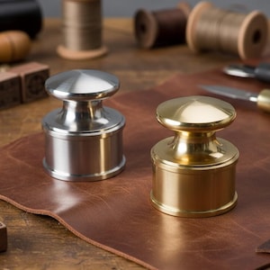 May include: Two metal leathercraft tools, one silver and one gold, sit on a brown leather surface. The tools have a cylindrical base and a rounded top. Other leatherworking tools, including spools of thread and knives, are in the background.