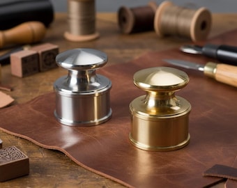 Könnte beinhalten: Zwei Lederhandwerkzeuge aus Metall, eines silberfarben und eines goldfarben, liegen auf einer braunen Lederoberfläche. Die Werkzeuge haben einen zylindrischen Sockel und eine abgerundete Oberseite. Weitere Lederwerkzeuge, darunter Garnrollen und Messer, sind im Hintergrund.
