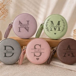 Op de afbeelding: Een verzameling gepersonaliseerde ronde meetlinten in verschillende kleuren, waaronder lavendel, groen, grijs, roze en bruin. Elk meetlint heeft een naam en initiaal, met een bijpassende polsband. De namen zijn Nancy, Madison, Denise, Sophia en Ashley.