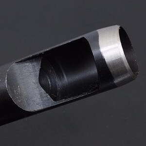 May include: A black metal punch tool with a silver-colored cutting edge. The tool is designed to punch holes in leather or other materials. The tool has a handle and a cylindrical body with a circular opening at the end.