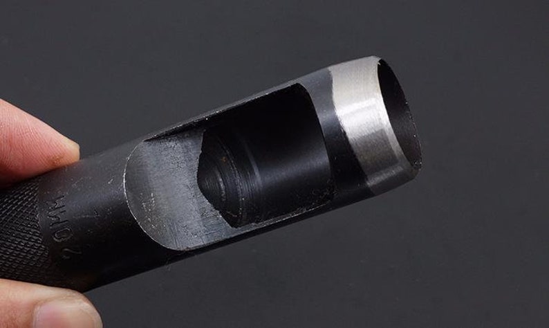 May include: A black metal punch tool with a silver-colored cutting edge. The tool is designed to punch holes in leather or other materials. The tool has a handle and a cylindrical body with a circular opening at the end.