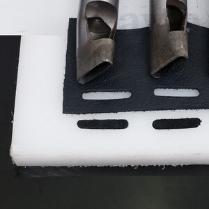 May include: Three metal leather hole punch tools on a black leather surface with a white cutting mat underneath.