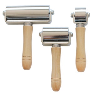 May include: Three metal and wood hand rollers of varying sizes are displayed against a white background. Each roller has a shiny, silver-colored cylindrical roller attached to a light-colored wooden handle. The handles are smooth and rounded.