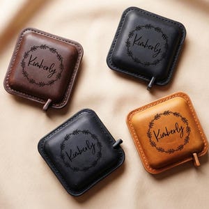 Può includere: Quattro sottobicchieri quadrati in pelle marrone, nera e beige. Ogni sottobicchiere presenta un design a ghirlanda circolare con il nome "Kimberly" al centro. Una piccola linguetta si estende da un lato di ogni sottobicchiere.