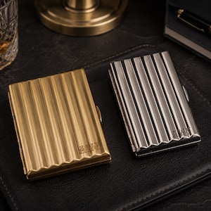 May include: Two metal cigarette cases. One is gold-toned, the other is silver-toned. Both have a ribbed design and are rectangular. The gold case has the initials "AWY" engraved on it. The silver case also has initials engraved.