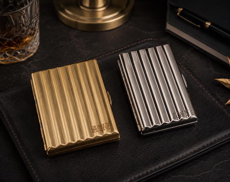 May include: Two metal cigarette cases. One is gold-toned, the other is silver-toned. Both have a ribbed design and are rectangular. The gold case has the initials "AWY" engraved on it. The silver case also has initials engraved.