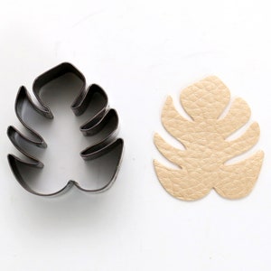 Może przedstawiać: Metalowa foremka do ciasteczek w kształcie liścia monstera obok skórzanego liścia monstera w kolorze beżowym.