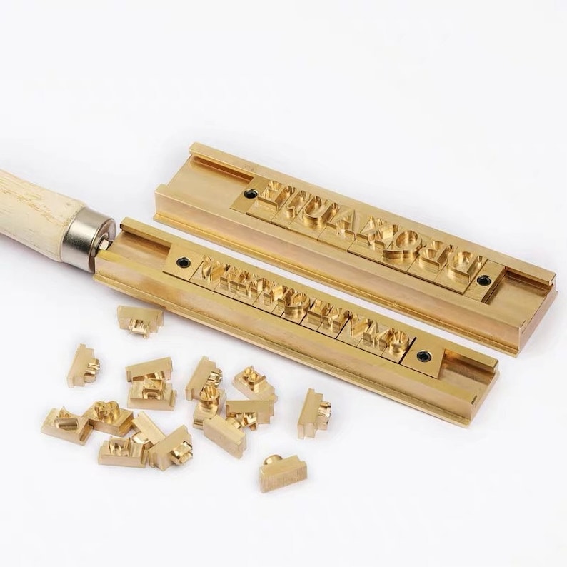 K&ouml;nnte beinhalten: Zwei Stempel-Sets aus Messing mit einem Holzgriff. Das obere Set hat die Buchstaben "PROUDLY" eingepr&auml;gt. Das untere Set hat eine Vielzahl von Buchstaben und Zahlen. Es gibt auch lose Messing-Stempel, die um die Sets herum verstreut sind.