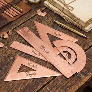 Puede incluir: Un conjunto de herramientas de dibujo de color cobre, que incluyen reglas, triángulos y un transportador, dispuestos sobre una superficie de madera. Las herramientas están grabadas con el nombre "Cayla". Otros elementos incluyen una brújula, lápices, un bolígrafo y una pila de cuadernos.