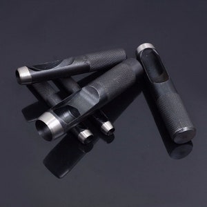 May include: A set of five black metal hole punch tools with different sized circular holes. The tools have a textured grip and are arranged on a black surface.