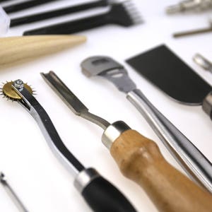 May include: Assortment of leatherworking tools, including a stitching groover, edge beveler, and a rotary wheel. The tools have wooden and metal handles, and are arranged on a white surface. The tools are used for crafting leather goods.