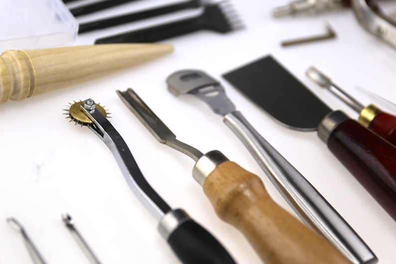 May include: Assortment of leatherworking tools, including a stitching groover, edge beveler, and a rotary wheel. The tools have wooden and metal handles, and are arranged on a white surface. The tools are used for crafting leather goods.